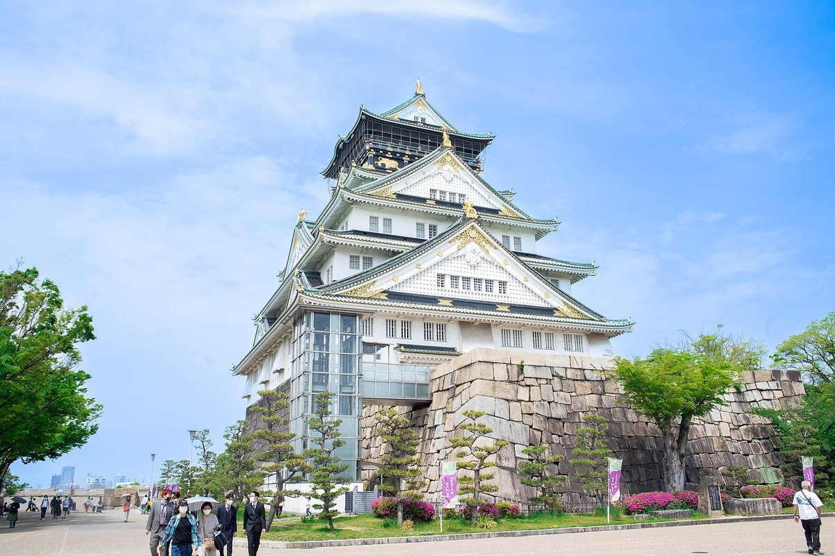 Osaka Castle main tower with white walls and green roofs above its stone base, with visitors walking in the park on a sunny spring day