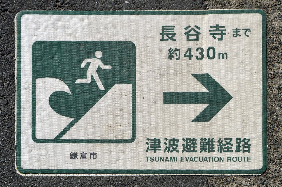 Green and white tsunami evacuation route sign set into the pavement in Kamakura, showing a running figure and an arrow toward higher ground