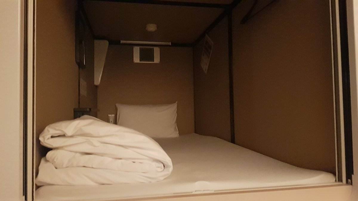 Inside a capsule hotel pod in Tokyo with a white mattress, pillow and folded duvet