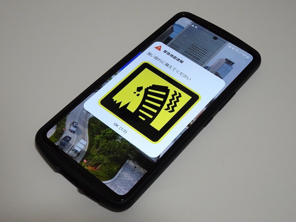 A smartphone screen showing a yellow Japanese Earthquake Early Warning alert with an icon of a shaking house