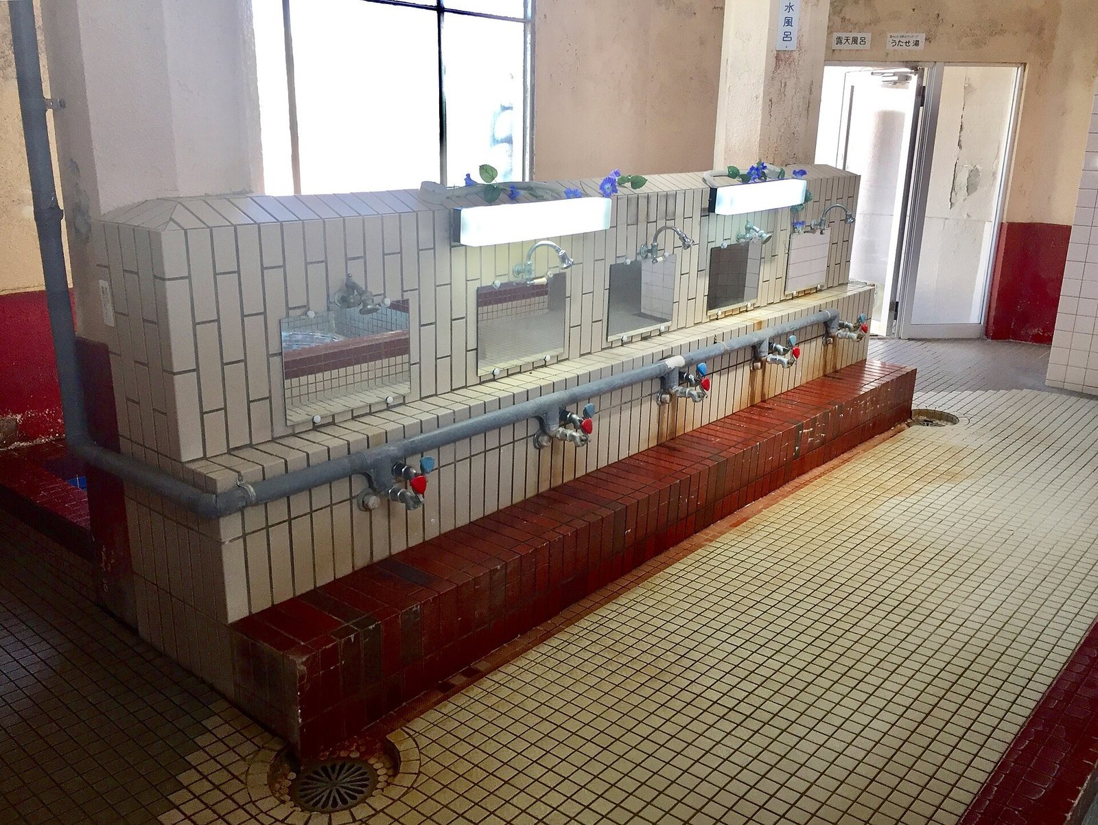 Row of low wash-station taps with mirrors and a tiled ledge in the bathing room of Imabari Radium Onsen, Ehime