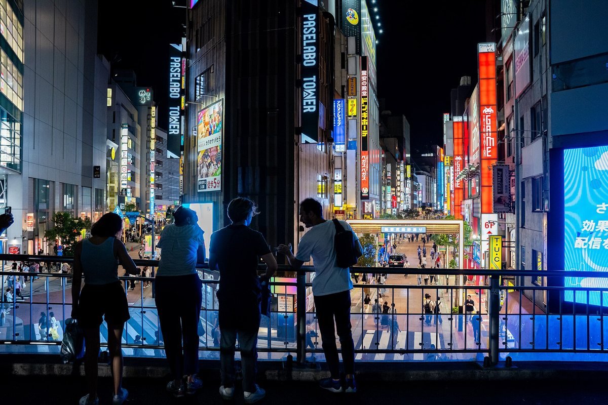 People standing at a railing above a busy Shinjuku street at night, with bright shop signs and lit-up buildings