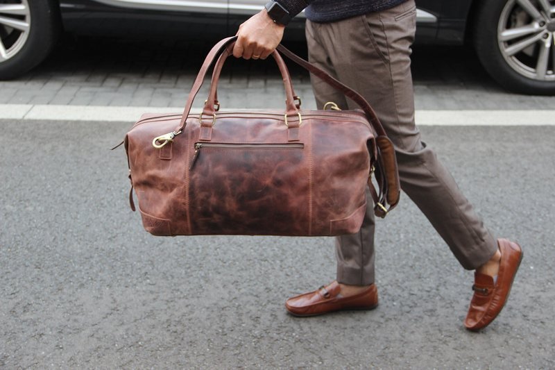 A person carrying a brown duffel-style holdall travel bag by its handles (item type photo, not the exact product)