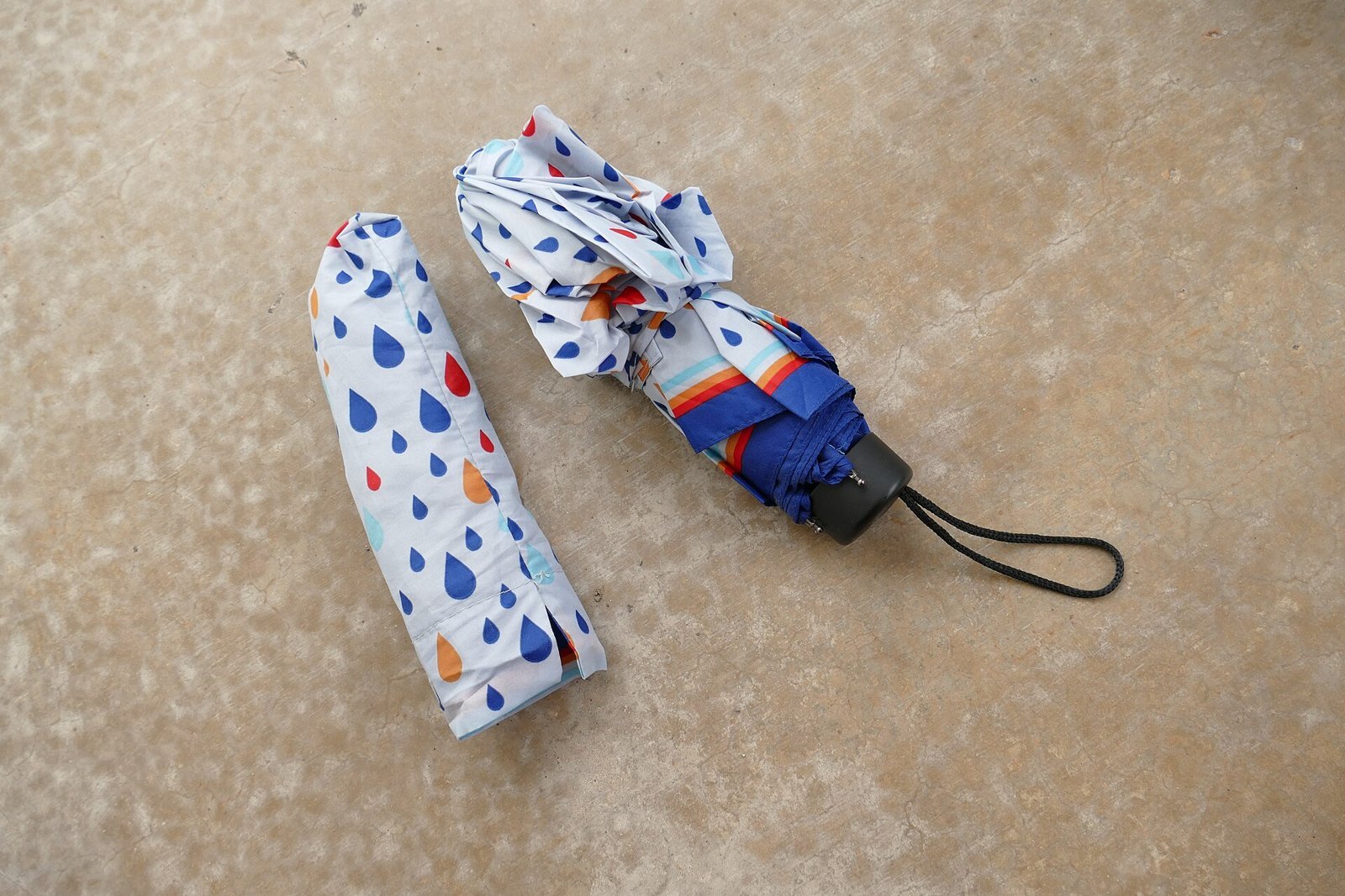 A small folding umbrella with a raindrop-print case lying on a stone floor (item type photo, not the exact product)